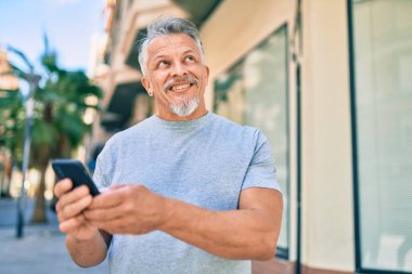 Orta yaşlı, ispanyol, kır saçlı, mutlu gülümseyen adam şehirde akıllı telefon kullanıyor..
