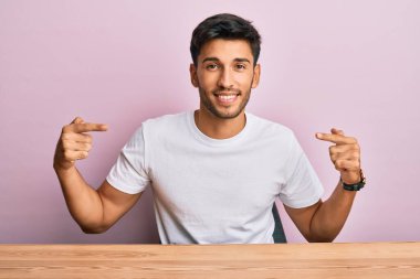 Masada oturan, rahat beyaz t-shirt giyen, kendine güvenen, yüzünde gülümseme olan, kendini gururlu ve mutlu gösteren genç yakışıklı bir adam.. 