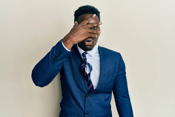 Handsome black man wearing business suit and tie peeking in shock covering face and eyes with hand, looking through fingers afraid