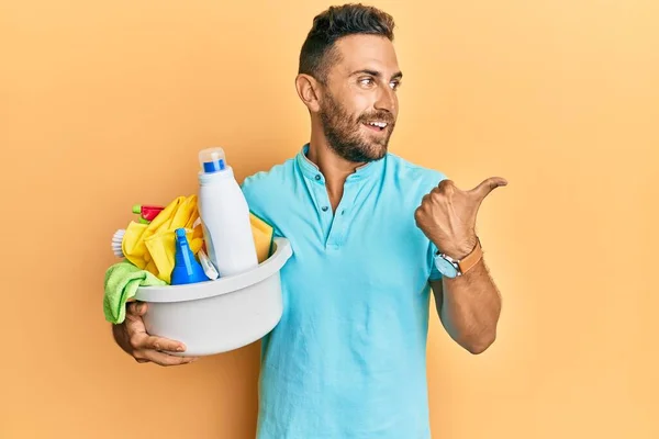 Handsome man with beard holding cleaning products pointing thumb up to the side smiling happy with open mouth