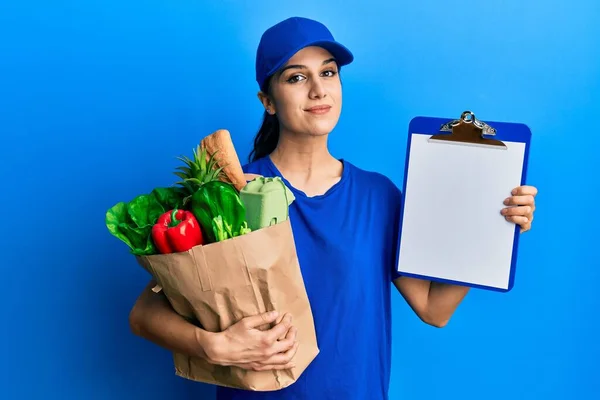 Young hispanic woman wearing courier uniform with groceries from supermarket and clipboard relaxed with serious expression on face. simple and natural looking at the camera.