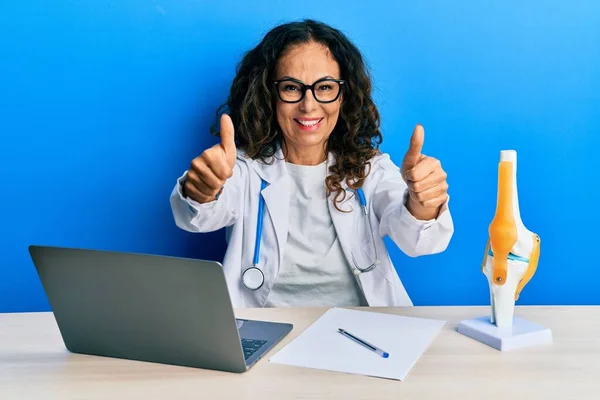 Beautiful middle age woman doctor at orthopedic clinic approving doing positive gesture with hand, thumbs up smiling and happy for success. winner gesture.