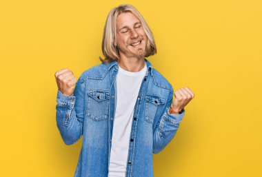 Sarışın, uzun saçlı, sıradan bir kot ceket giyen, kolları havada, gülümseyen ve başarı çığlıkları atan, çok mutlu ve heyecanlı beyaz bir adam. kutlama konsepti. 