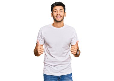 Gündelik beyaz tişört giyen genç, yakışıklı bir adam elinde olumlu bir jest yapıyor, gülümsüyor ve mutlu. Neşeli ifade ve kazanan jest. 