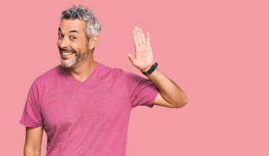 Orta yaşlı, gri saçlı, sıradan kıyafetler giyen, mutlu ve gülümseyen, dostça bir jest yapan bir adam. 