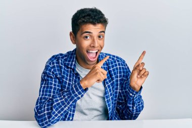 Genç, yakışıklı, Afro-Amerikan bir adam, gündelik giysiler içinde masaya oturmuş gülümsüyor ve kameraya bakıyor. İki eliyle ve parmaklarıyla işaret ediyor.. 
