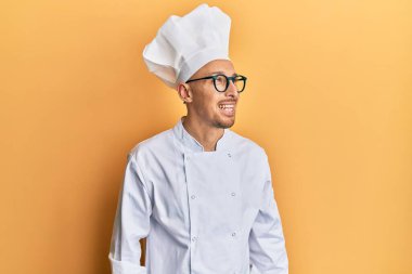 Profesyonel aşçı üniforması giyen kel bir adam yüzünde gülümsemeyle yan yana bakıyor, doğal bir ifade. Kendinden emin gülen. 