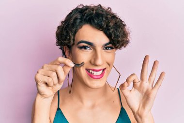 Takma kirpik tutan genç adam makyaj yapıyor. Parmaklarıyla imza atıyor, gülümsüyor. Mükemmel bir sembol. 