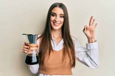 Güzel esmer, elinde çözünür kahve makinesi olan genç bir kadın parmaklarıyla imza atıyor, gülümseyerek mükemmel bir sembol oluşturuyor. 