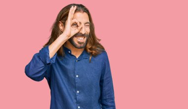 Uzun saçlı ve sakallı çekici bir adam. Günlük kıyafetler giyiyor. El hareketleriyle gülümsüyor.. 