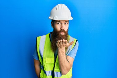 Uzun sakallı, güvenlik kaskı takan ve yansıtıcı ceketli kızıl saçlı bir adam kameraya bakıp havada bir öpücük savuruyor. Güzel ve seksi. aşk ifadesi. 