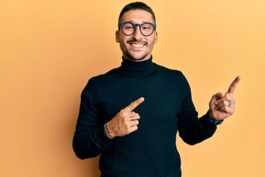 Balıkçı yaka kazak giyen, gözlüklü, gülümseyen ve kameraya bakan yakışıklı bir adam.. 