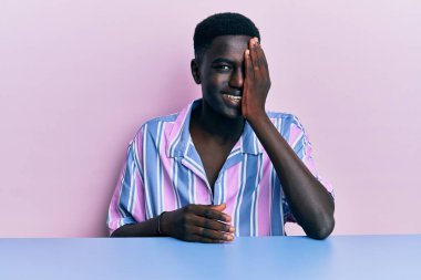 Genç, Afro-Amerikan bir adam, gündelik giysiler içinde masada oturuyor, bir gözünü eliyle kapatıyor, yüzünde kendine güvenen bir gülümseme ve sürpriz bir duygu var.. 