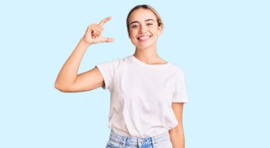 Genç, güzel, sarışın, günlük beyaz tişörtler giyen, gülümseyen, kendine güvenen, el hareketi yapan, parmaklarıyla işaret eden ve kameraya bakan bir kadın. ölçüm kavramı. 