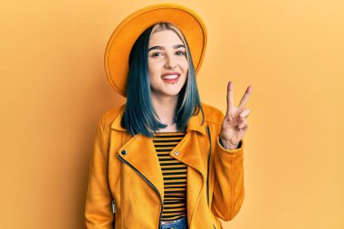 Sarı şapka ve deri ceket giyen, kameraya gülümseyen ve parmaklarıyla zafer işareti yapan genç, modern bir kız. İki numara.. 