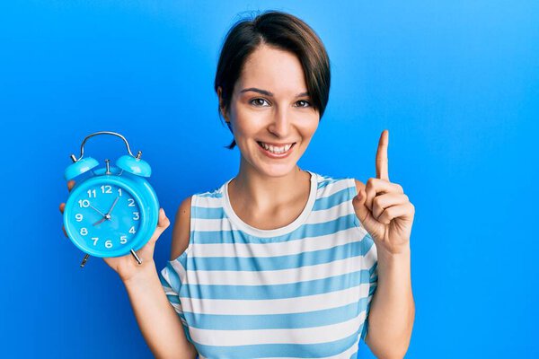Young brunette woman with short hair holding alarm clock smiling with an idea or question pointing finger with happy face, number one 