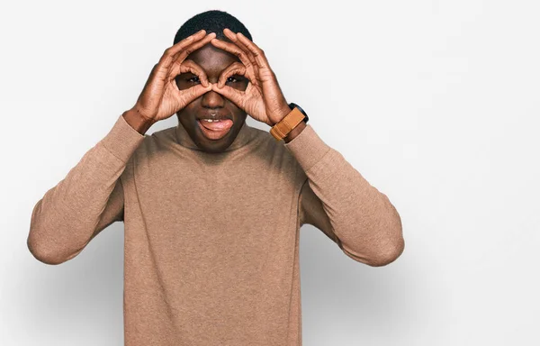 冬のカジュアルなセーターを着た若いアフリカ系アメリカ人の男性は 双眼鏡が舌を突き出しているようなOkジェスチャーをして 目は指を見ています クレイジーな表現 — ストック写真