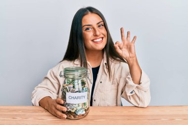 Güzel İspanyol bir kadın elinde para dolu bir kavanoz tutuyor parmaklarıyla imza atıyor, gülümsüyor ve mükemmel bir sembol oluşturuyor. 