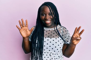 Saçları örgülü genç siyahi bir kadın sekiz numarayı gösterip gösterirken kendinden emin ve mutlu bir şekilde gülümsüyor.. 