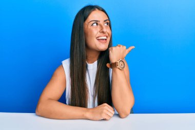 Güzel İspanyol kadın, gündelik giysiler içinde masaya oturmuş başparmağını kaldırıyor ve ağzı açık gülümsüyor. 