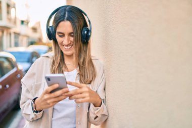 Beyaz kadın şehirde akıllı telefon ve kulaklık kullanarak mutlu bir şekilde gülümsüyor..