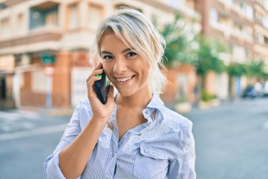 Genç sarışın kadın gülümsüyor. Şehirdeki akıllı telefondan konuşuyor..