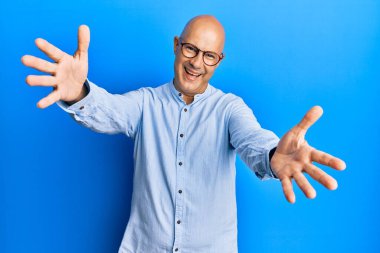 Orta yaşlı, kel bir adam, günlük kıyafetler ve gözlük takmış kameraya bakıyor ve sarılmak için kollarını açmış gülümsüyor. Mutluluğu kucaklayan neşeli bir ifade.. 