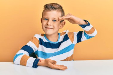 Sevimli beyaz çocuk, sıradan kıyafetler giyip masaya oturmuş büyük ve büyük boyut işareti, ölçü sembolü göstererek el kol hareketi yapıyor. Kameraya bakıp gülümsüyor. ölçüm kavramı. 