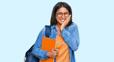 Öğrenci çantası takan, elinde kitap tutan, gülen, utanan, ağzı ellerle kapatan, dedikodu ve skandal konsepti olan genç Latin kız. 