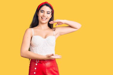 Zarif görünümlü esmer bir kız. Elleriyle büyük ve büyük bir işaret gösteriyor. Ölçü sembolü. Kameraya bakıp gülümsüyor. ölçüm kavramı. 