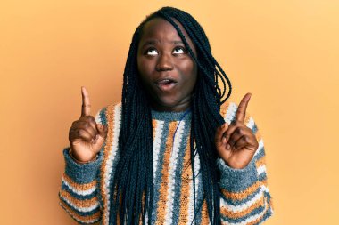 Kış süveteri giyen genç siyah kadın şaşırmış ve şaşırmış bir şekilde yukarı bakıp parmaklarını göstermiş ve kollarını kaldırmıştı.. 