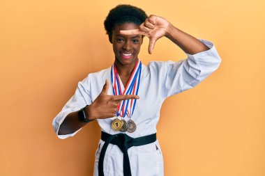 Genç Afrikalı Amerikalı kız karate kimonosu ve siyah kuşak takmış gülümsüyor ve elleri ve parmakları gülen yüzlü bir çerçeve yapıyor. yaratıcılık ve fotoğrafçılık kavramı. 