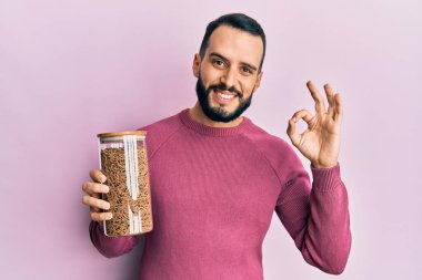 Sakallı genç adam sağlıklı tahıl gevreği kavanozunu tutuyor. Parmaklarıyla işaret veriyor, gülümsüyor. Mükemmel bir sembol. 