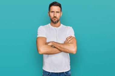 Sakallı, sıradan, beyaz tişörtlü, kuşkucu ve gergin, yüzünde çapraz kollu, onaylamayan bir ifade olan yakışıklı bir adam. negatif kişi. 