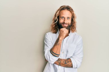 Sakallı, uzun saçlı, sıradan beyaz gömlekli, kameraya kolları çapraz ve çenesi açık gülümseyen yakışıklı bir adam. Olumlu düşünmek. 