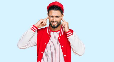 Sakallı genç adam beysbol üniforması giyiyor. Kulaklarını parmaklarıyla kapatıyor. Gürültülü müzik için rahatsız edici bir ifade takınıyor. sağır kavramı. 