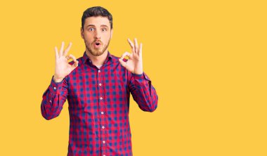 Normal gömlekli yakışıklı genç adam. Şaşırmış ve şaşırmış görünüyor. Onaylama sembolünü parmaklarıyla yapıyor. çılgın ifade 