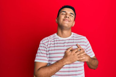 Gündelik kıyafetler giyen genç Arap adam elleri göğsünde gülümsüyor, gözleri kapalı ve yüzünde minnet ifadesi var. sağlık kavramı. 