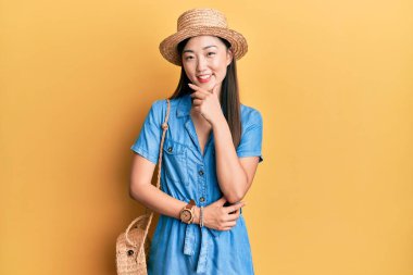 Yazlık şapka takan genç Çinli kadın kameraya kendinden emin bakıyor kollarını kavuşturup gülümsüyor ve çenesini kaldırıyor. Olumlu düşünmek. 