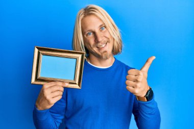 Kafkas asıllı, uzun saçlı, boş afiş tutan mutlu ve pozitif gülümseyen, başparmağıyla mükemmel ve onay işareti yapan genç bir adam. 