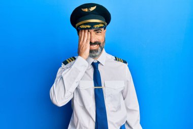 Orta yaşlı, sakallı, gri saçlı, uçak pilotu üniforması giyen, bir gözünü elinle kapatan, yüzünde kendine güvenen bir gülümseme ve şaşırtıcı bir duygu olan bir adam.. 