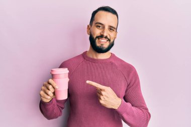 Sakallı genç adam bir fincan kahve içerken mutlu bir şekilde elini ve parmağını göstererek gülümsüyor. 