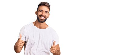 Latin asıllı genç adam, günlük beyaz tişört giyiyor. Başarı işareti yapıyor. Elle olumlu jest yapıyor. Neşeli ifade ve kazanan jest. 