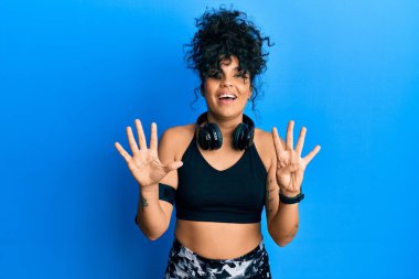 Spor kıyafetleri giyen ve kulaklık takan genç İspanyol kadın dokuz numarayı gösterip işaret ederken kendinden emin ve mutlu bir şekilde gülümsüyor.. 