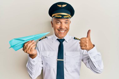 Yakışıklı, orta yaşlı, uçak pilotu üniforması giyen, elinde kağıt uçak tutan mutlu ve pozitif gülümseyen, başparmağıyla mükemmel ve onay işareti yapan olgun bir adam. 