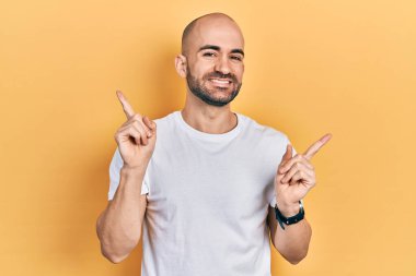 Gündelik beyaz tişört giyen genç ve kel bir adam farklı yönleri gösteren kendine güvenen bir şekilde gülümsüyor. reklam için kopyalama alanı 