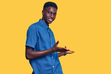 Genç, Afro-Amerikan bir adam, günlük giysiler içinde gülümseyerek içeri girmeye davet ediyor. 