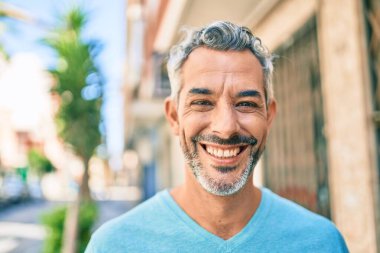 Orta yaşlı, kır saçlı, gülümseyen bir adam şehrin caddesinde mutlu bir şekilde yürüyor..