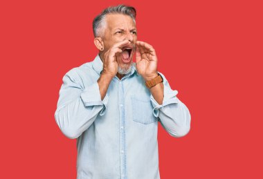 Orta yaşlı, gri saçlı, günlük kıyafetler giyen, elleri ağzında kızgın bir şekilde bağıran bir adam. 