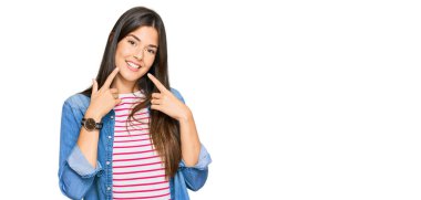 Genç esmer kadın gündelik kıyafetler giyip neşeli bir şekilde gülümsüyor ve parmaklarıyla ve ağzıyla işaret ediyor. diş sağlığı kavramı. 